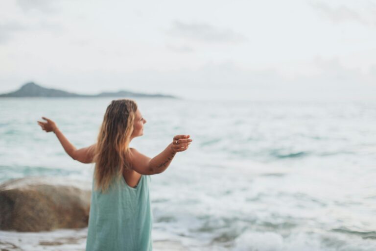 Side view of happy female traveler in dress enjoying summer day on ocean beach and recreating with outstretched arms