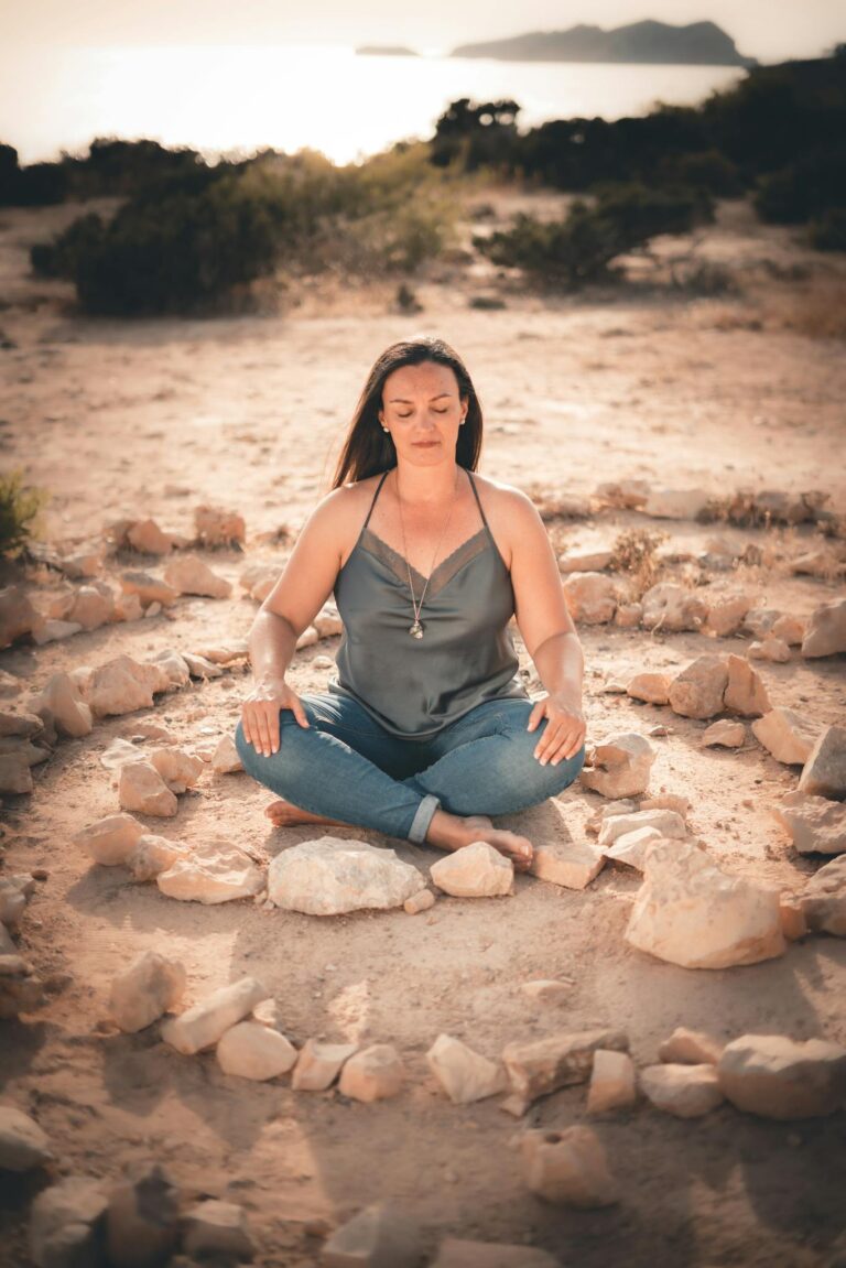 Woman meditating cross-legged on a sandy beach surrounded by stones, captured at sunset.
