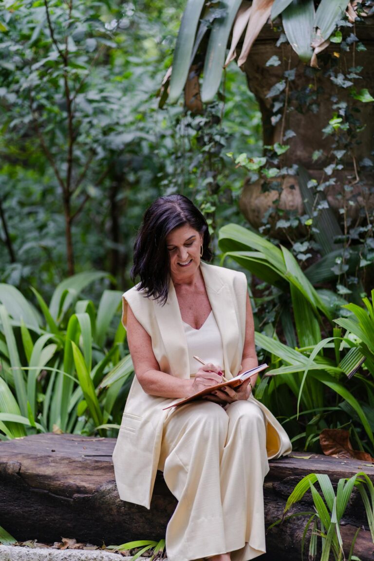 Woman sitting on a log writing in a notebook amidst lush greenery, conveying creativity and tranquility.